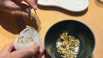 Hands placing gold leaf into mold, table surface.