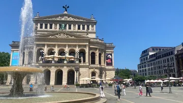 Opernhaus mit Brunnen, Menschen spazieren vorbei.