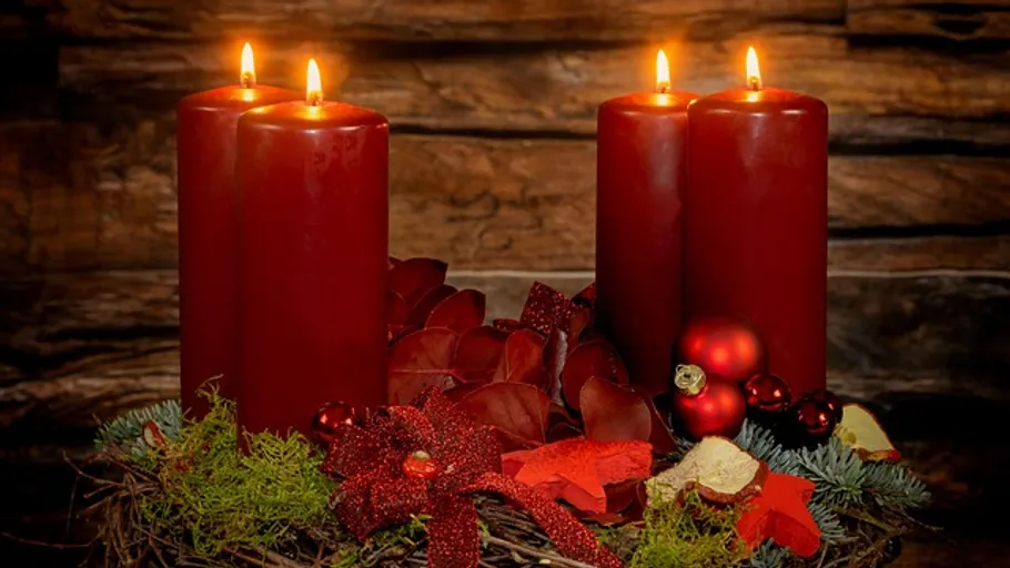 Four lit red candles on wreath, wooden background.